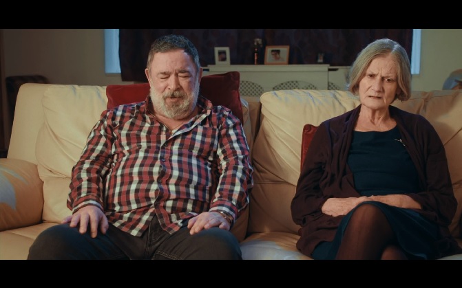 An elderly couple sitting on a cream couch in their front room, talking to someone off-screen.