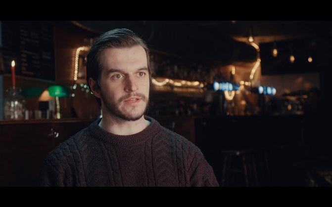 Bearded man in a dark trendy bar setting wearing a thick cable knit jumper talking to someone off-screen.