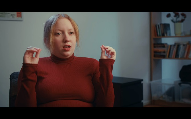 Young blonde woman in a burgundy turtle neck talking to someone off-screen.